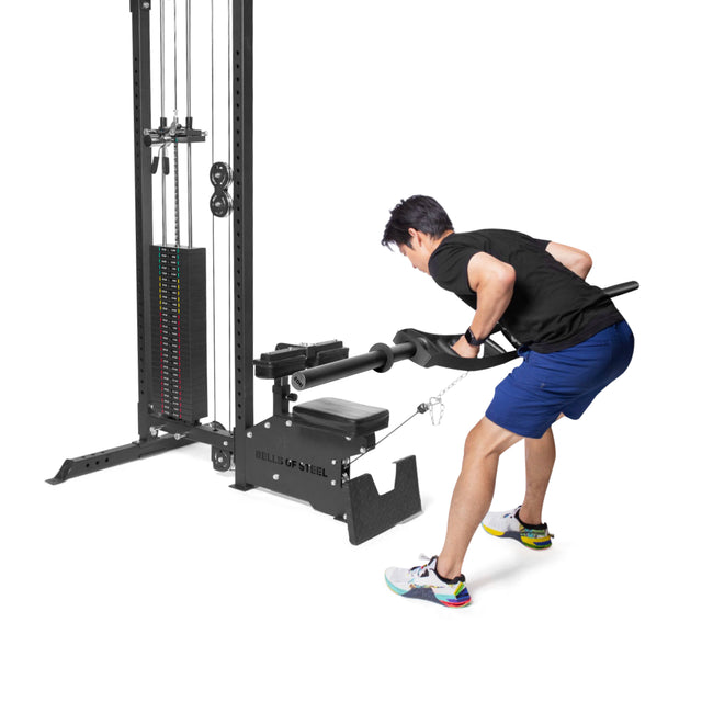 A person in a black shirt and blue shorts uses the Bells of Steel Arch Nemesis Swiss Bar on a cable machine to do bent-over rows, pulling the handle attached to weights and engaging their back muscles against a plain white background.