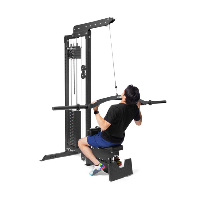 A person in a black shirt and blue shorts performs a lat pulldown on a sleek cable machine using Bells of Steel's Arch Nemesis Swiss Bar, expertly pulling the wide bar as the specialty barbells engage smoothly with the pulley system.