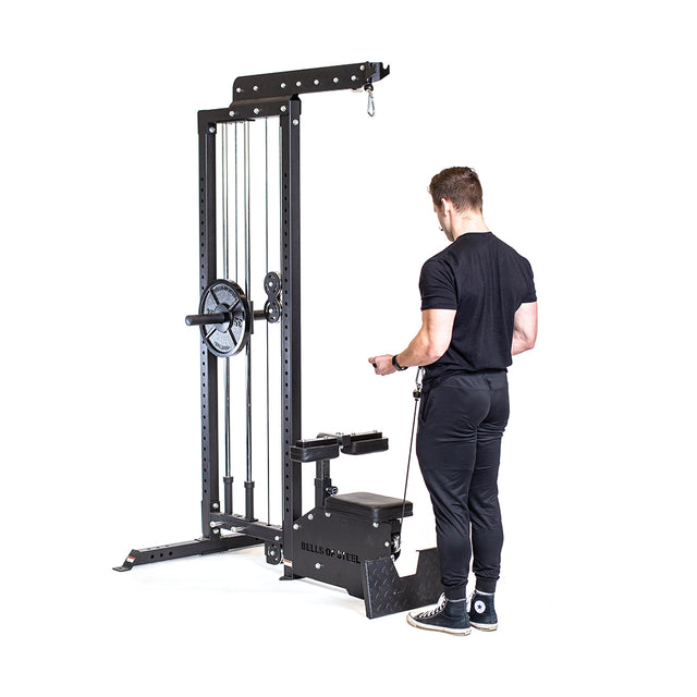 Dressed in black, an individual uses the Bells of Steel Lat Pulldown Low Row Machine. The black equipment with weights and pulleys enhances their workout as they grip the handle with both hands, performing an exercise similar to a lat pulldown while facing the machine.