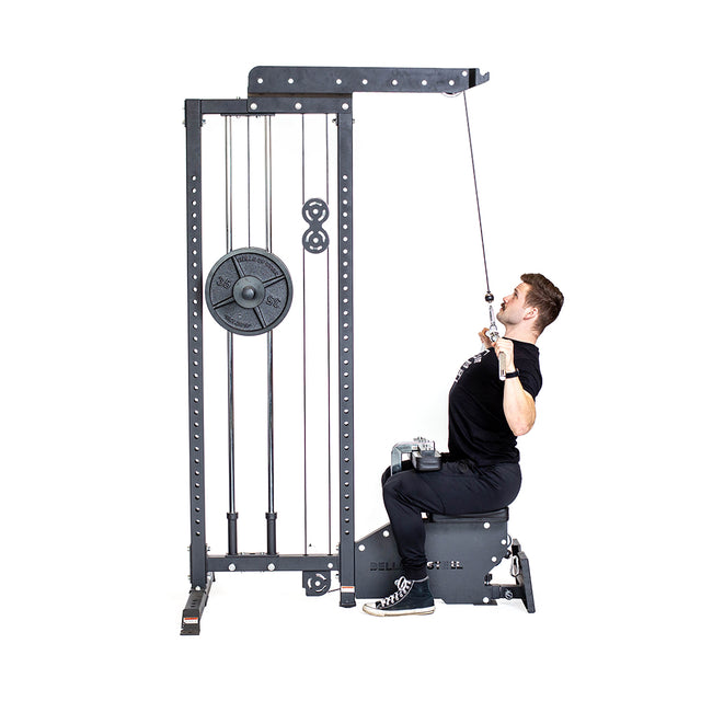 A man in workout attire uses the Bells of Steel Lat Pulldown Low Row Machine in his home gym. He grips the bar, pulling it down toward his chest. The machine features multiple weight plates and a pulley system.