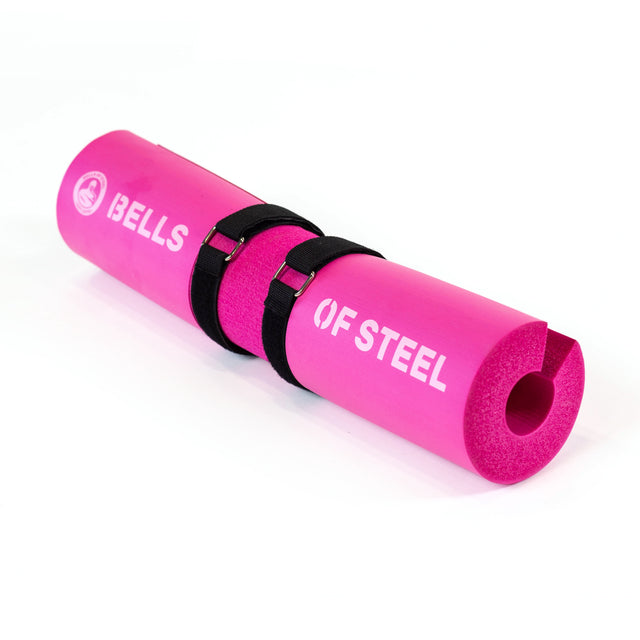 A plush pink foam Bells of Steel barbell pad with black straps, featuring an ergonomic cylindrical design for comfort during weightlifting.