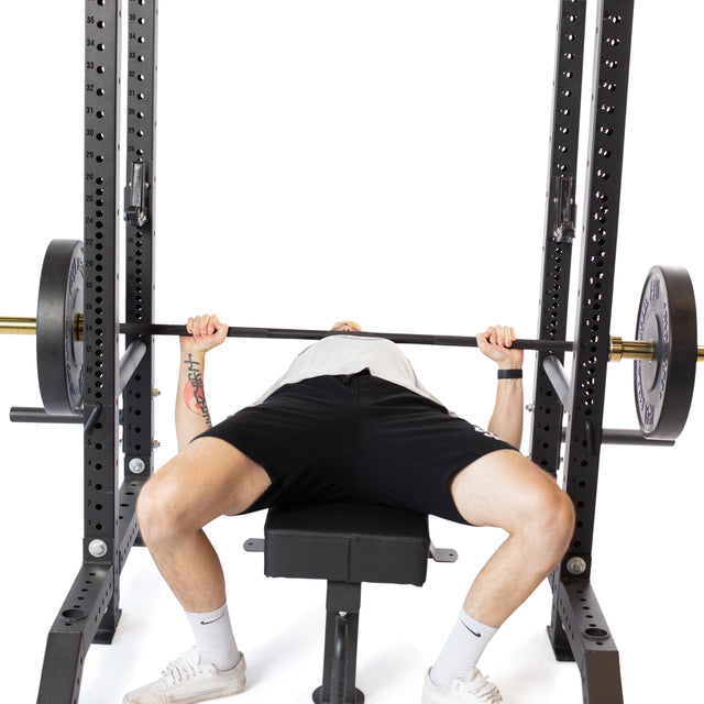 A person is bench pressing a barbell in a metal power rack using Pin Pipe Safeties from Bells of Steel. Dressed in a white shirt, black shorts, and white socks, they ensure safe training with precise movements and equipment.