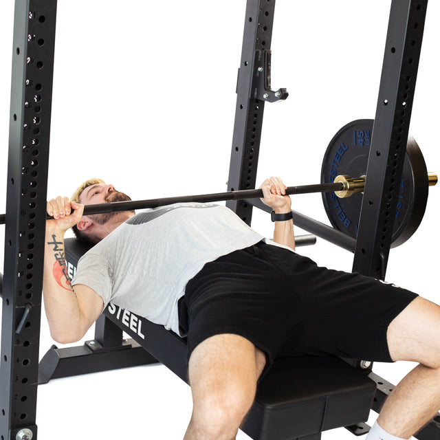 A person lies on a bench press setup inside a power rack, lifting a barbell. Wearing a gray shirt and black shorts, they focus intently upward. For added safety during their workout, they rely on Bells of Steel's Pin Pipe Safeties with weights balanced on both sides.