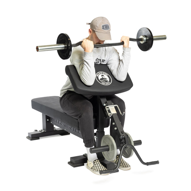 Equipped with Bells of Steel Bench Attachments, a person in a gray cap, long-sleeve shirt, and checkered shoes sits intently performing barbell preacher curls using the arm rest and weighted bar effortlessly.