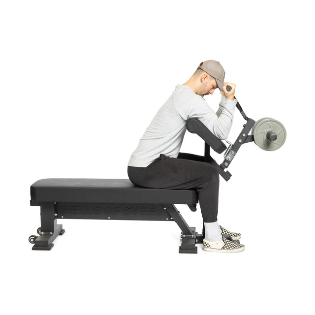 A man in a gray shirt and hat uses Bells of Steel Bench Attachments. He sits focused on the bench, legs straight for extensions, wearing black pants and checkered shoes.