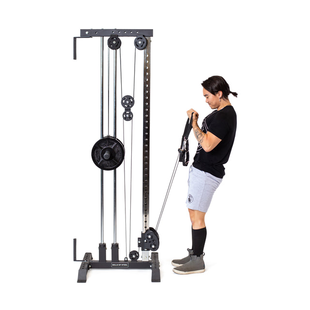 Wearing a black shirt and gray shorts, a person intently works out on the Bells of Steel Cable Tower, gripping its adjustable handles. The freestanding equipment features a visible weight stack and pulleys to enhance their fitness routine.