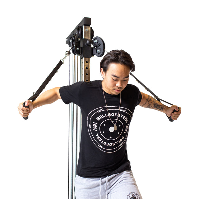 A person in a gray t-shirt and pants is using the Bells of Steel Cable Tower for a chest exercise, intensely pulling the adjustable handles outward with both hands.