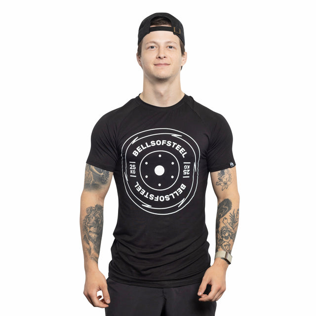 A person in a black Bells of Steel Bamboo T-Shirt and a backward cap stands facing forward, displaying tattooed arms, against a white background.