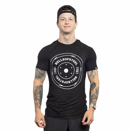 A person in a backward black cap and a buttery soft Bells of Steel Bamboo T-Shirt with a kettlebell graphic and weight markings smiles at the camera. The shirt, reading "Bells of Steel," pairs perfectly with their black shorts, showcasing tattooed arms.