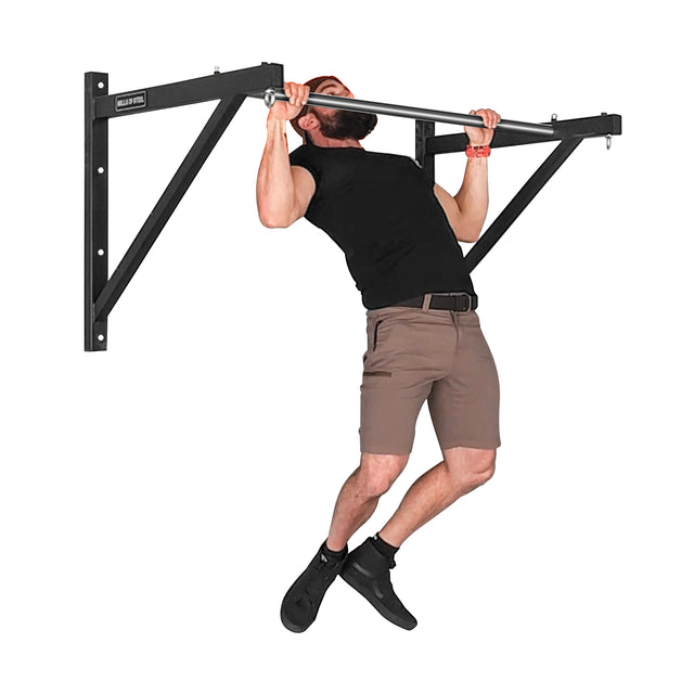 A bearded man intensely performs a pull-up on the Bells of Steel Adjustable Wall or Ceiling Mounted Pull Up Bar. Dressed in a black t-shirt, brown shorts, and black shoes, he showcases his dedication to fitness with this kipping-friendly equipment during his indoor routine.