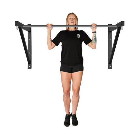 A tattooed person performs a pull-up on the Bells of Steel Adjustable Wall or Ceiling Mounted Pull Up Bar, wearing a black T-shirt and shorts. Their focused gaze and form stand out against a plain white background.