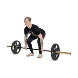 A tattooed person in a black T-shirt and pants stands against a plain white background, preparing to lift a barbell with ergonomic handles and Bells of Steel's Rubber Coated Iron Weight Plates, while wearing white sneakers.