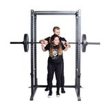 A woman squats with a barbell inside the Bells of Steel Residential Power Rack (2.3" x 2.3", ⅝" holes) with j-cups, as a man spots behind her; both wear black athletic gear—ideal for any home gym setting.
