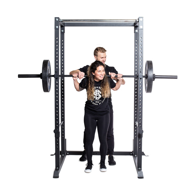 A woman squats with a barbell inside the Bells of Steel Residential Power Rack (2.3" x 2.3", ⅝" holes) with j-cups, as a man spots behind her; both wear black athletic gear—ideal for any home gym setting.