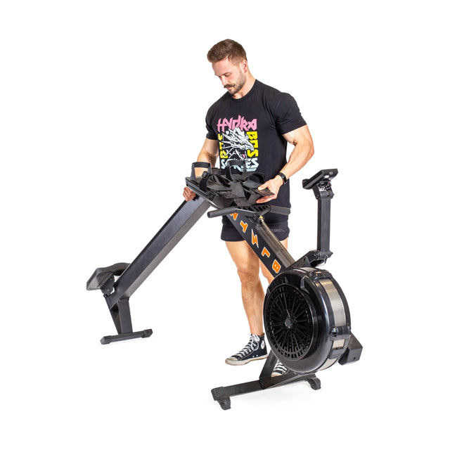 A person in a black T-shirt and shorts, wearing sneakers, is assembling the Blitz Air Rower by Bells of Steel. This black machine, perfect for low-impact cardio workouts, features a circular fan at the front and adjustable footrests, offering a full-body workout experience.