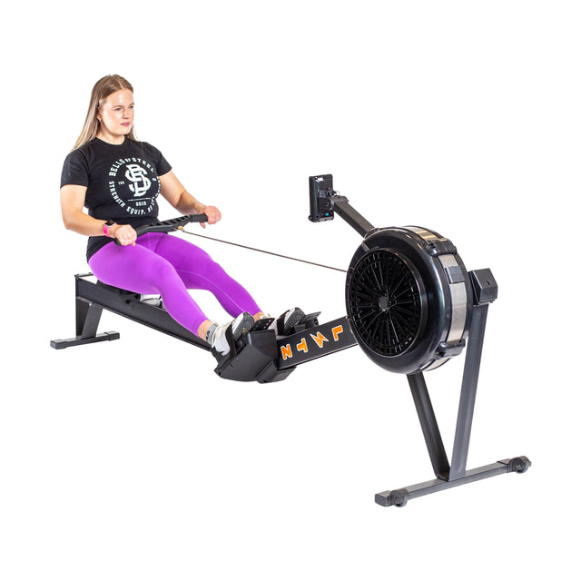 A woman in a black t-shirt and purple leggings uses the Blitz Air Rower from Bells of Steel, ideal for low-impact cardio. She pulls the handle with both hands, seated with her feet secure on the footrests against a plain white background.