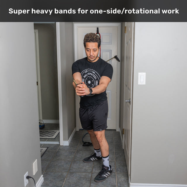 A man stands in a hallway, holding Bells of Steel USA Ripcords Premium Resistance Bands with both hands, performing a rotational exercise. Text above reads: “Super heavy bands for one-side/rotational work.”.