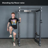A person stands next to a squat rack, lifting one knee with a Bells of Steel USA Human Trainer - Leg Cords resistance band around his ankle. The text “Standing hip flexor raise” is shown at the top.