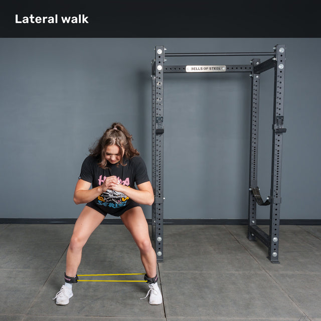 Wearing athletic gear, a woman demonstrates a lateral walk using the Bells of Steel USA Human Trainer - Leg Cords around her legs in a gym, positioned in front of a squat rack.