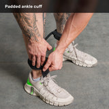 A tattooed individual secures a black padded ankle cuff from the Human Trainer - Leg Cords by Bells of Steel USA around their lower leg, preparing for a leg and glute workout while wearing white sneakers with green accents on a gray floor.