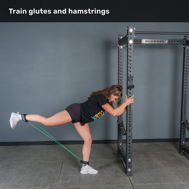 A person uses the Bells of Steel USA Human Trainer - Leg Cords attached to a metal rack, performing a kickback exercise that targets the glutes and hamstrings. Leaning forward and holding onto the rack with both hands, they extend one leg back for focused muscle activation.