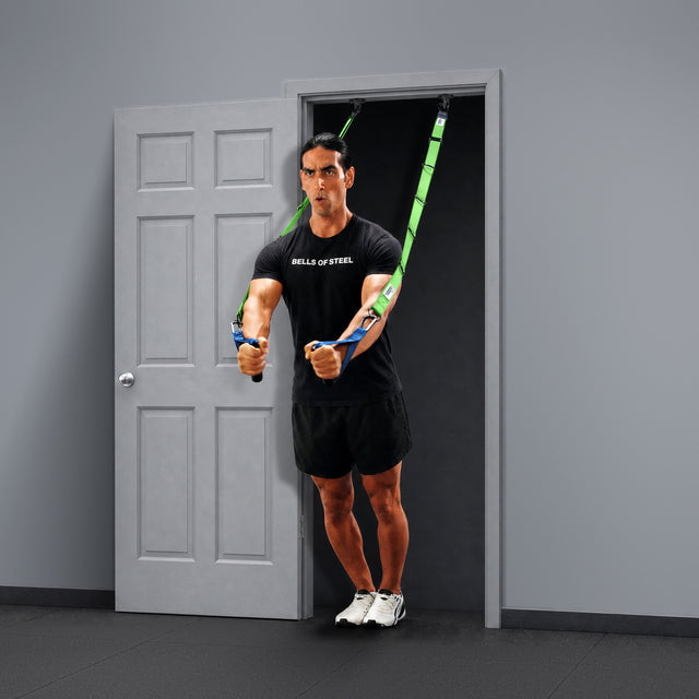 Wearing a black t-shirt and shorts, a person works out in a minimalist room, leaning forward and gripping the handles of green Human Trainer - Ceiling Mounts by Bells of Steel USA, which are securely fixed overhead for suspension training.