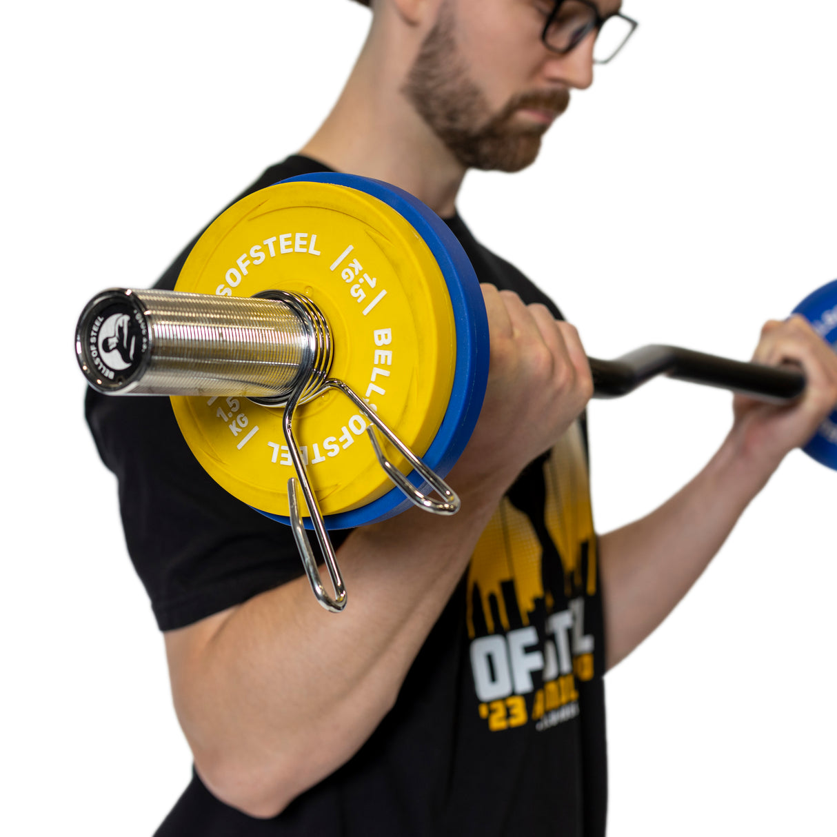 Sporting a black shirt with yellow and white graphics, a person in glasses focuses intently while lifting a barbell. The blue and yellow weight plates are secured by Bells of Steel's Spring Collars against the plain white background.