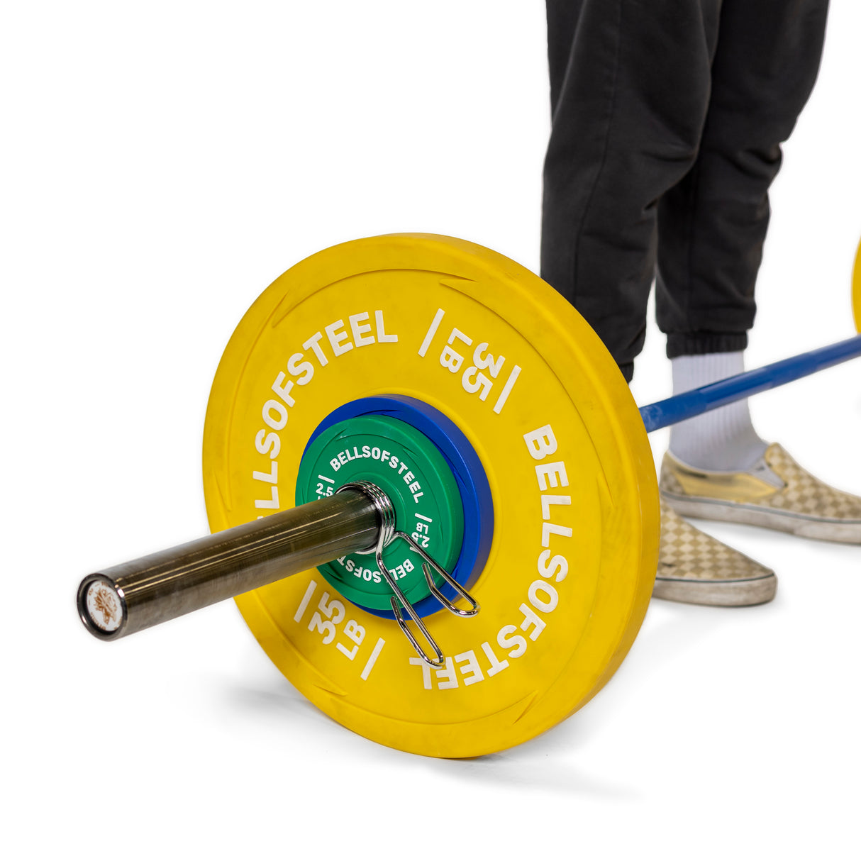 A person in checkered slip-on shoes and black pants adjusts Spring Collars by Bells of Steel on a barbell with Olympic sleeves and polished chrome, loaded with yellow and green weight plates. Only their lower legs are visible, ensuring the plates are secure.