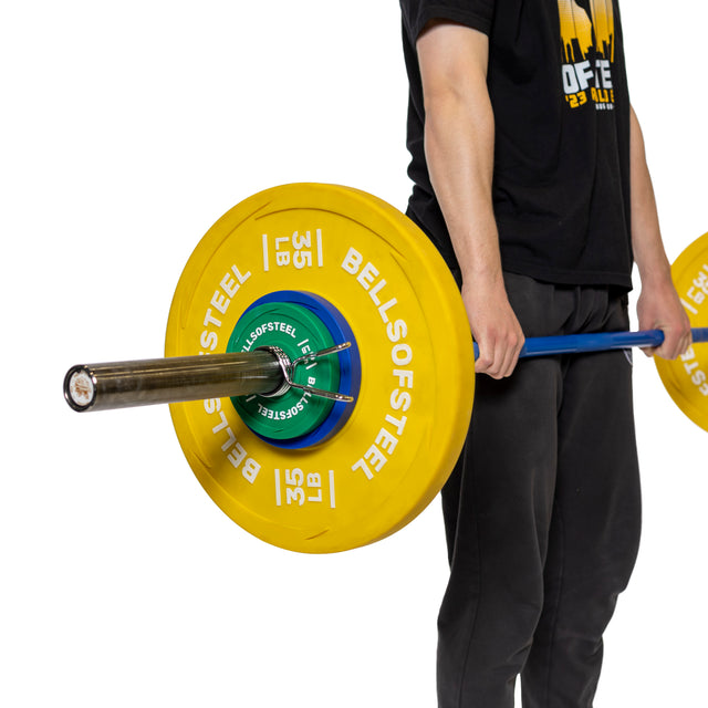 A person in black holds a barbell with large yellow 35 lb "Bells of Steel" weight plates and Bells of Steel Spring Collars at thigh height. The background is white.