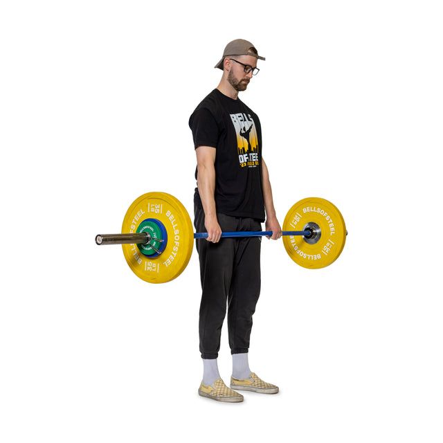 A man in a cap, glasses, black t-shirt and pants, and checkered shoes stands holding a barbell with yellow weight plates secured by Bells of Steel Spring Collars, arms straight down, against a white background.