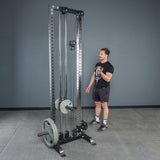 A person stands next to a tall, black cable pulley machine loaded with weight plates, using the Bells of Steel Fat Bar - Single D Handle to improve grip strength in a gym with gray walls and a concrete floor.