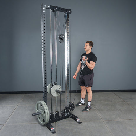 A person stands next to a tall, black cable pulley machine loaded with weight plates, using the Bells of Steel Fat Bar - Single D Handle to improve grip strength in a gym with gray walls and a concrete floor.