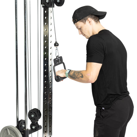 Wearing a black shirt and cap, a person uses the cable machine at the gym. Tattoos on their arm enhance grip strength as they focus intently, gripping the Bells of Steel Fat Bar - Single D Handle attached to the machine.