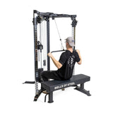 A person in a black shirt and gray cap uses the Bells of Steel All-in-One Trainer for a seated row on its bench, featuring adjustable pulleys and weights, demonstrating its versatility.