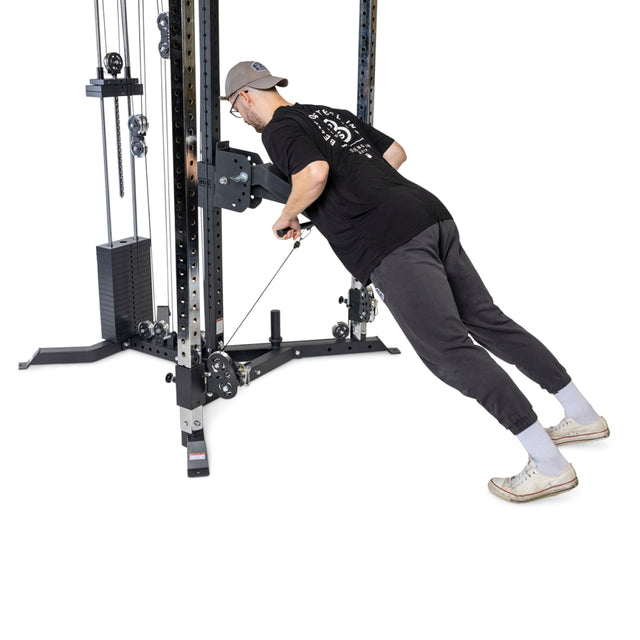 Clad in a cap, glasses, and casual attire, an individual works out on the Bells of Steel All-in-One Trainer cable machine, leaning forward to grip the handles with weights stacked on this multifunctional gear.