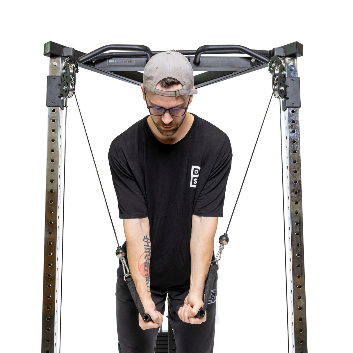 A person in a black shirt and gray cap exercises with the Bells of Steel All-in-One Trainer, pulling the cable crossover handles with focused effort against a plain white background.