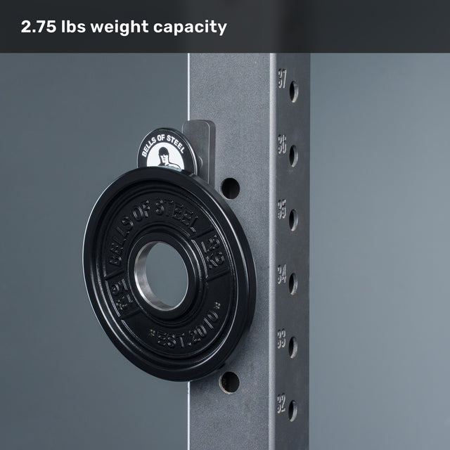 A black 2.75 lbs weight plate is mounted on a metal rack with numbered adjustment holes, next to the Bells of Steel USA Selfie Sentinel – Magnetic Rack Phone Holder, a compact gym tripod with a secure mount for convenient workout tracking. At the top, text reads “2.75 lbs weight capacity” on a gray background.