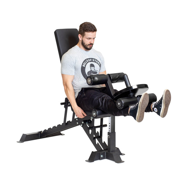 A person wearing a gray shirt and black pants uses the Buzz-Saw Heavy-Duty Adjustable Bench by Bells of Steel in a gym, pushing against padded bars with both legs. The bench features an adjustable back pad and a black cushioned seat, set on a sturdy metal frame for heavy-duty use.