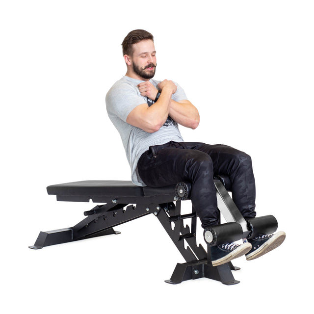 A man works out on a Bells of Steel Buzz-Saw Heavy-Duty Adjustable Bench, which has a 1,000lb capacity. He wears a gray t-shirt, black pants, and sneakers while sitting with arms crossed. The bench features an adjustable back pad against a plain white background.