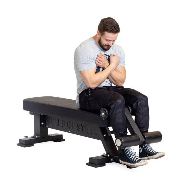 A man in a gray T-shirt and black pants sits on a Bells of Steel workout bench with Bench Attachments. His arms are crossed, feet on padded leg supports, wearing black sneakers with white soles.