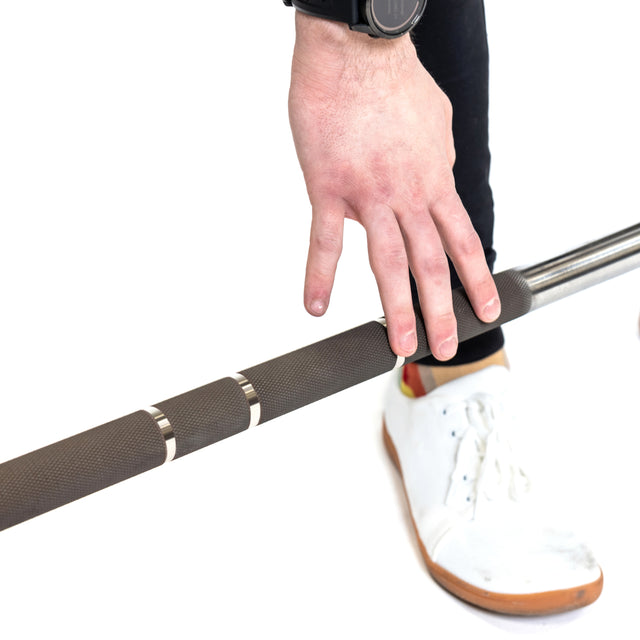 A person grips the textured handle of a Bells of Steel Hydra Seven Ring Bar, readying for a lift. They wear a black watch, black pants, and white sneakers accented with red and yellow, against a clean white backdrop that emphasizes their dedication to powerlifting.