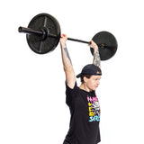 A person in a black cap and graphic tee lifts the Bells of Steel Short Utility Barbell overhead against a plain white background—ideal inspiration for your home gym setup.