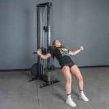 A woman in athletic wear uses the Bells of Steel USA Aluminum Handles Pair with Bearings on a cable machine, leaning back with arms extended, standing on a gray floor against a plain gray wall.