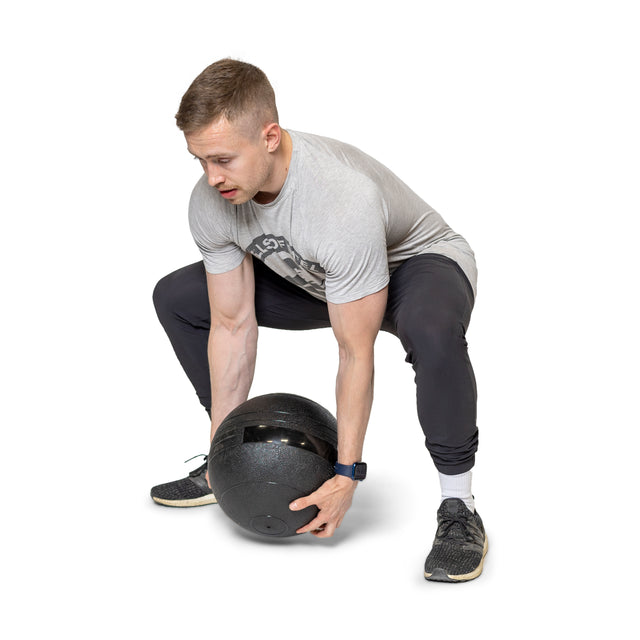 A person in a gray t-shirt and black pants is powerfully squatting with a large Bells of Steel Slam Ball. Wearing a smartwatch and black sneakers, they are perfectly equipped for intense cardio workouts against a plain white backdrop that highlights their focus and strength.