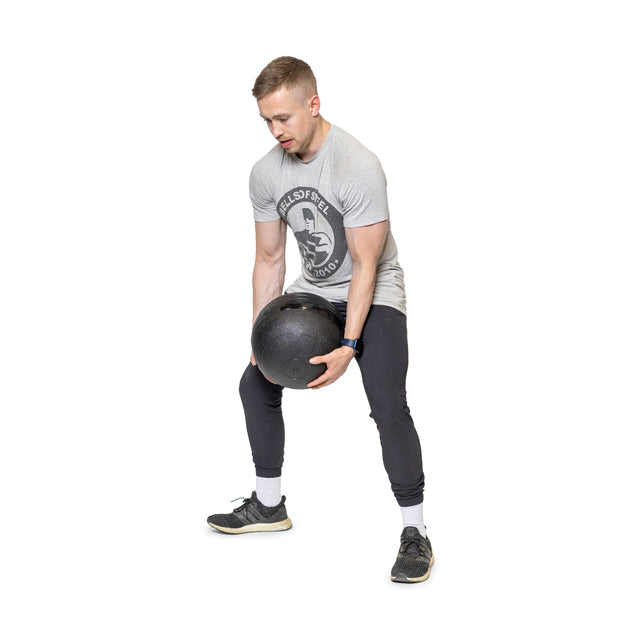 Dressed in a gray t-shirt and dark pants, a man focuses intensely as he harnesses explosive power by lifting heavy black Bells of Steel Slam Balls with a wide stance, adding intensity to his cardio workouts.