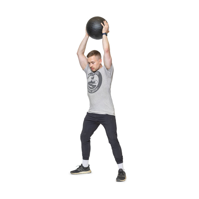 A man in a gray T-shirt and black pants shows explosive power by lifting a black medicine ball overhead, legs apart, focused and strong on a white background, embodying Bells of Steel's Slam Balls in intense cardio workouts.