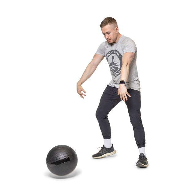 The person in Bells of Steel athletic wear is performing a cardio workout using Slam Balls, showcasing explosive power. They are positioned with knees slightly bent and arms outstretched, deeply focused on their routine against a white background.