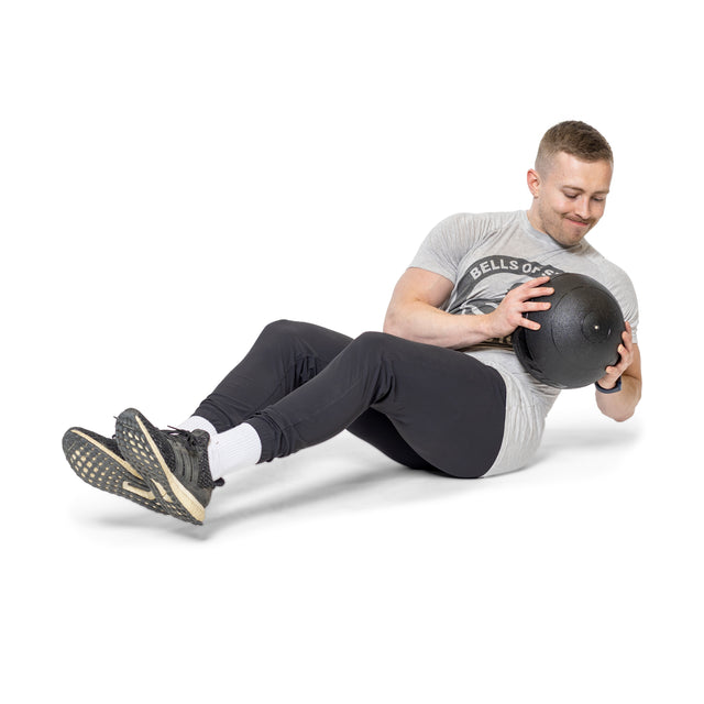 A man exercises with a Bells of Steel Slam Ball, performing the Russian twist. Seated on the floor, knees bent, feet slightly elevated, he sports a gray t-shirt, black pants, and black athletic shoes against a plain white backdrop.