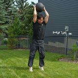 A person dressed in black athletic wear stands on grass, holding a Bells of Steel Slam Ball overhead and preparing to slam it down, demonstrating explosive power. In the background are trees, a fenced yard, and a dark building.