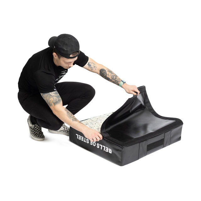 A tattooed person in black attire and cap squats, peeling back the "Bells of Steel" deadlift pad replacement cover, revealing its speckled interior. Made of durable 600 Denier Polyester material, it's perfect for sturdy deadlift pads.